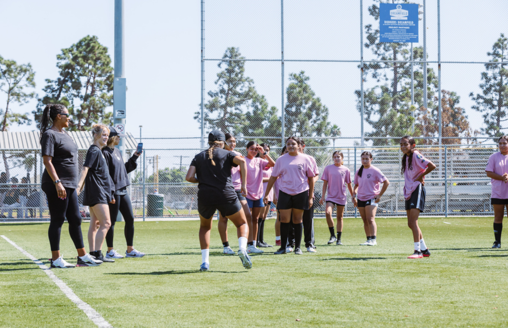 A group of adolescent girls listen to a coach giving instructions on a soccer field.
